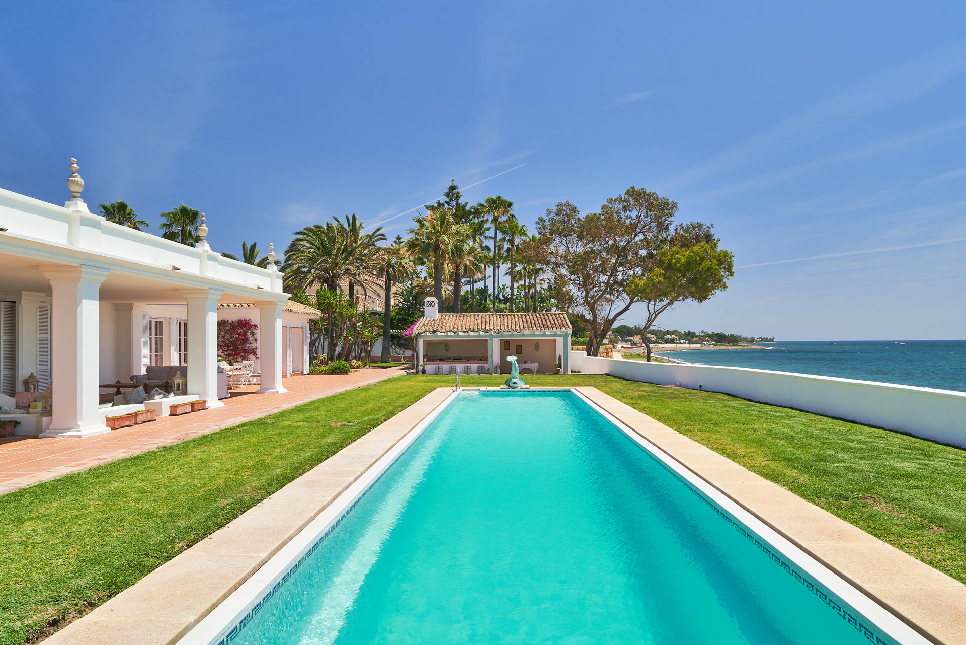 Imagen de una piscina en primer plano, con panorama del mar y vista lateral de una casa y una construccion en ultimo plano.